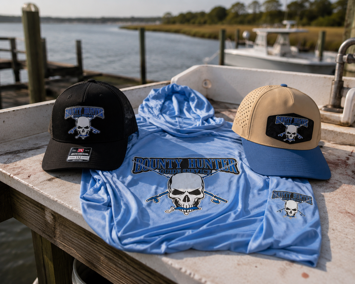 Blue t-shirt with skull design, black cap, and tan and blue cap on a dock by water.